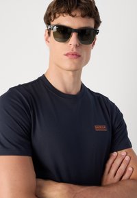 Tricou bleumarin cu guler rotund și mâneci scurte, dotat cu un mic logo portocaliu. Ochelari de soare negri cu lentile întunecate și accente metalice.