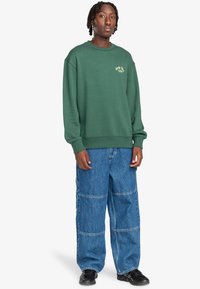 Felpa verde con polsini e scollatura a coste, caratterizzata da un piccolo logo bianco. Abbinata a pantaloni in denim azzurro chiaro con gamba ampia e cuciture.