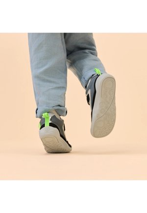 Persoon die lichtblauwe spijkerbroek en grijze sneakers met neon groene trekbandjes draagt, van achteren getoond terwijl hij met een halve stap beweegt op een beige achtergrond.