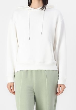 Mujer con sudadera blanca lisa con cordones y pantalones verde claro, de pie contra un fondo blanco.