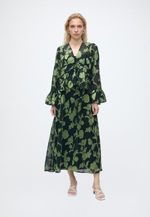 Femme portant une longue robe noire transparente avec des motifs floraux verts et des manches à volants, debout avec une expression neutre et des sandales blanches à brides.