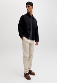 Bărbat purtând o jachetă neagră cu fermuar, cămașă închisă la culoare, pantaloni bej și pantofi maro din piele, stând cu o mână în buzunar pe un fundal simplu.