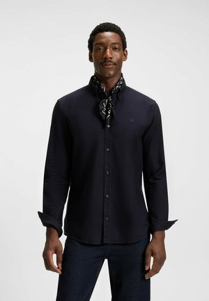 Navy long-sleeve shirt with button-down collar, featuring a small logo, paired with a patterned black and white scarf around the neck.