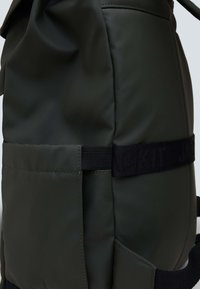 Zaino impermeabile verde con finitura opaca, spallacci regolabili e una prominente fascia nera con il marchio. Dettagli della tasca laterale visibili.