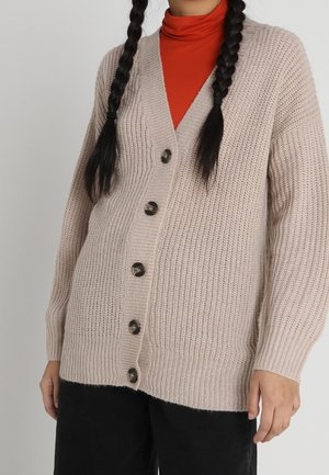 Personne portant un cardigan beige en grosse maille avec cinq boutons sur un col roulé orange et un pantalon noir, avec deux longues tresses.