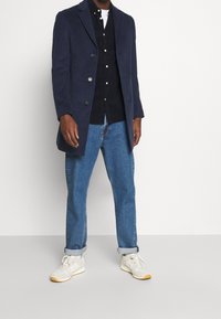 Cappotto in lana blu navy con vestibilità su misura, design doppiopetto, camicia nera con bottoni e jeans azzurri. Calzature: sneakers crema con dettagli color sabbia.