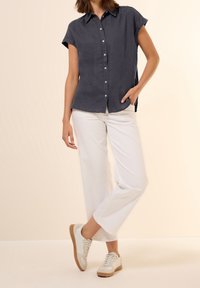 Granatowa koszula z krótkim rękawem i kołnierzykiem typu spread, w połączeniu z białymi, szerokimi spodniami i jasnymi trampkami. Gładka tekstura materiału.