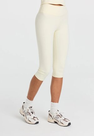 Underkrop af en person iført cremefarvede capri-leggings, hvide crew-sokker og hvide og grå New Balance-sportsko.