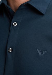 Tamno plava majica polo sa oštrim kragnom, srebrnim dugmadi i reljefnim logotipom orla na lijevoj strani prsiju. Glatka tekstura tkanine.