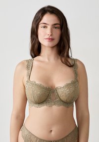 Koronkowy biustonosz w ziemistym odcieniu zieleni, z falbaniastymi krawędziami i dziurką z przodu. Regulowane ramiączka uzupełniają delikatny kwiatowy wzór.