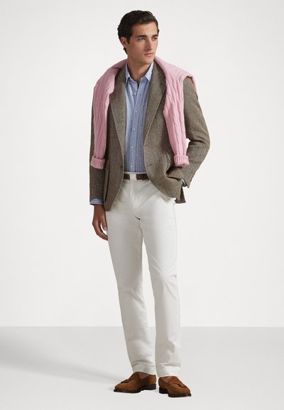 Brūns heringa kaula žakets, gaiši zils svītrains krekls, balti bikses un rozā adīts džemperis, kas uzkarsēts pār pleciem; brūnas loafers.