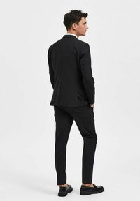 Svart skreddersydd dress med slim fit. Har spiss krage og stramme bukser. Bært med svarte sko, sett fra baksiden. Glatt tekstur.
