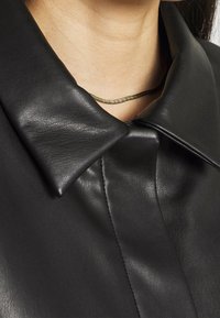 Camicia in pelle nera con ampio colletto, texture liscia e cuciture pulite, con una sottile collana metallica indossata sotto.