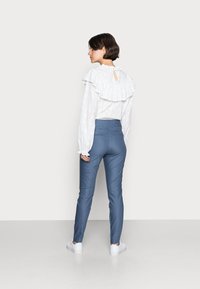 Hvit langermet topp med volangkrage, kombinert med blå tettsittende bukser; hvite sneakers synlige, minimalistisk bakgrunn.