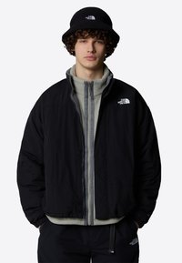 Sort zip-up jakke med grå foring over en lys grå fleece, parret med en matchende sort bucket hat med et logo og mørke bukser.