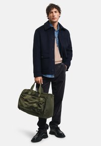 Marineblauer Wollmantel über beigem Pullover, blauer Hemdbluse, dunklen Hosen. Olivgrüne Reisetasche mit zwei Griffen. Schwarze Schuhe. Minimalistisches Design.