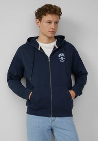 Felpa con zip color navy, dotata di cordoncino, tasche a marsupio e dettaglio del logo sul lato sinistro del petto. Realizzata in tessuto morbido; con polsini e fondo a coste.