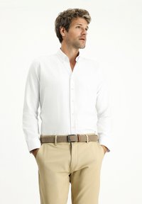 Camicia bianca con texture liscia, abbinata a pantaloni beige chiaro e una cintura intrecciata. Caratterizzata da un colletto classico e maniche lunghe.