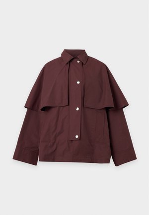 Femme portant une cape bordeaux, une chemise rose, un pantalon beige à jambes larges, des ballerines noires, tenant un grand sac à main noir sur un fond uni.