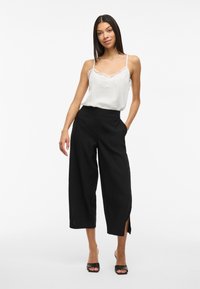 Camiseta de tirantes con encaje blanco, combinada con pantalones de pierna ancha negros de cintura alta con aberturas laterales. Modelo usando tacones negros de punta abierta.