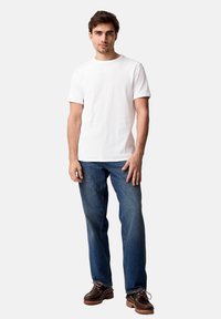 Tricou alb cu guler rotund, blugi din denim albastru și pantofi maro cu șireturi, având o talpă texturată. Modelul stă drept, cu mâinile pe laturi, având o expresie neutră.