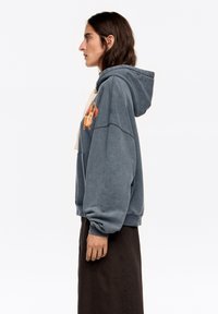 Niebiesko-szara bluza z kapturem, ozdobiona haftowanym wzorem psa, wykonana z przytulnego materiału, z prążkowanymi mankietami. Połączona z długą, ciemną spódnicą.