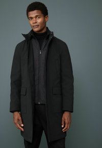 Parka negra resistente al agua con forro interior con cremallera, dos bolsillos frontales y un cuello alto. Textura suave y diseño elegante.