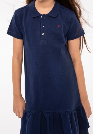 Dunkelblaues Polokleid mit Kragen, kurzen Ärmeln und gerafftem Rüschensaum. Mit kleinem roten besticktem Logo auf der Brust.
