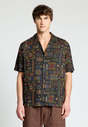 Giovane uomo che indossa una camicia nera a maniche corte con bottoni e motivi geometrici e tribali colorati, abbinata a pantaloni marroni.