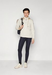 Felpa con cappuccio beige chiaro con zip e coulisse, abbinata a leggings blu navy con dettagli a toppa. Il modello indossa scarpe sportive bianche e porta uno zaino.