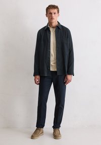 Dunkelblaues Hemd über einem cremefarbenen Pullover, kombiniert mit dunkler Hose und beigen Wildlederschuhen. Hintergrund ist schlicht und neutral.