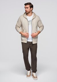 Felpa beige con zip e cordini, abbinata a una maglietta bianca e pantaloni cargo marroni con zip laterali. Sneakers marroni e crema.