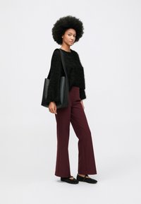 Afro frizurás nő fekete bolyhos pulóvert, bordó nadrágot, fekete tűsarkú lapos cipőt visel, és egy nagy fekete válltáskát hord.