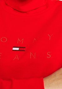 Sudadera de cuello alto de algodón rojo con "TOMMY" y "JEANS" bordados en dorado, que presenta una pequeña franja de logotipo horizontal en rojo, blanco y negro.