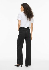 Plati juoda kelnės iš lygios medžiagos su kišenėmis gale, derinamos su paprasta balta marškinėliais. Modelis stovi šonu.