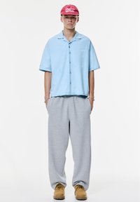 Cămașă bleu deschis cu mânecă scurtă și nasturi, pantaloni de trening gri și cizme de lucru bej. Șapcă roșie cu text alb care include detalii cu logo.