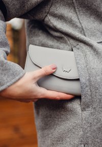 Light gray leather clutch with a curved flap design, stitched edges, and a small silver decorative accent, held against a gray coat.