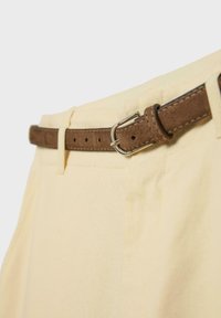 Pantalon beige avec une ceinture en suède marron dotée d'une boucle argentée passant à travers les passants sur un fond clair.