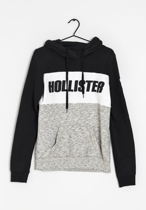 La sudadera con capucha presenta una parte superior negra, una banda blanca con el texto "HOLLISTER" y una sección inferior texturizada gris. Incluye un bolsillo delantero.
