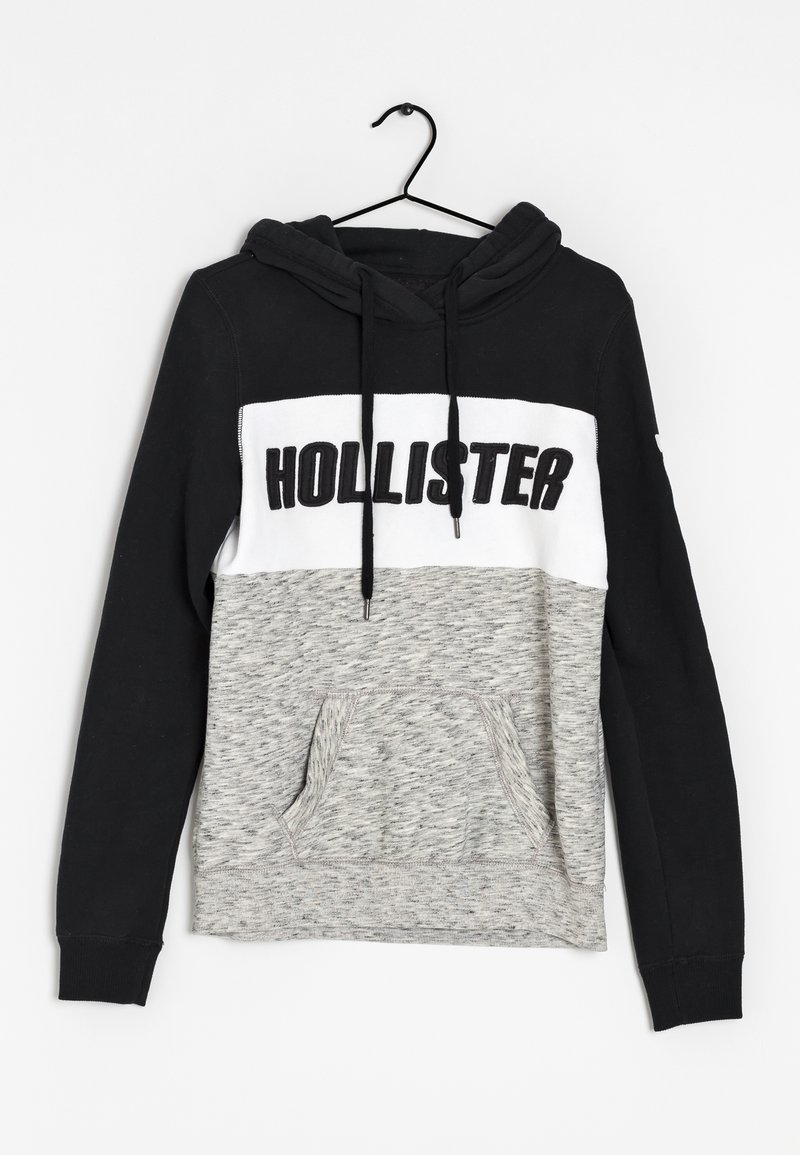 La sudadera con capucha presenta una parte superior negra, una banda blanca con el texto "HOLLISTER" y una sección inferior texturizada gris. Incluye un bolsillo delantero.