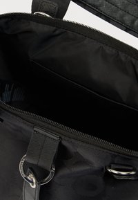 Μαύρη υφασμάτινη τσάντα με φερμουάρ, ανοιχτή για να δείξει μια άδεια εσωτερική τσέπη και μεταλλικές λεπτομέρειες με κρίκο στα λουριά της λαβής.