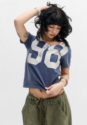 Jonge vrouw met zwart haar tilt een blauw shirt met "56" op, toont tatoeages en draagt groene broek met trekkoord, armbanden en piercings.