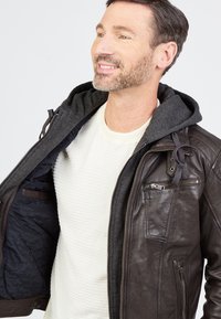 Ruskea nahkatakki, jossa on kiinnitetty harmaa huppari, vetoketjullinen etuosa, rintatasku ja sininen tikattu vuori. Valkoinen paita alla.