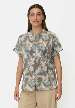 Vrouw met lang bruin haar draagt een kort mouw knoophemd met blauwe, bruine en beige abstracte patronen en een beige broek, staand tegen een witte achtergrond.