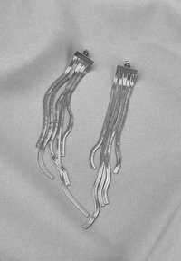 Transparente Acrylohrringe mit verlängerten, gewellten Strängen, akzentuiert mit metallischen Clips, ruhend auf einem strukturierten grauen Hintergrund.