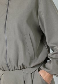 Graue, leichte Jacke mit Reißverschluss vorne, elastischem Bund und Seitentaschen. Verfügt über eine glatte Textur und einfache Designlinien.