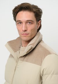 Beżowa puchowa kurtka z kołnierzem z sztruksu. Kurtka ma gładki, wyściełany design z zapięciem na zamek oraz teksturowymi akcentami.