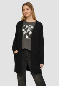 Zwarte lange cardigan van zacht breimateriaal, met een open voorkant, geribbelde manchetten en twee voorkanten zakken. Gecombineerd met een grijze graphic T-shirt.