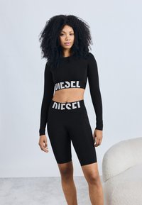 Czarny zestaw składający się z długiego rękawa crop top i krótkich spodenek z wysokim stanem, z białym logo "DIESEL" na pasie oraz gładką, elastyczną tkaniną.