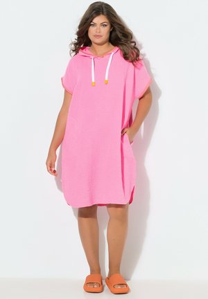 Kobieta z długimi brązowymi włosami, ubrana w luźną różową sukienkę z kapturem i kieszeniami oraz pomarańczowe klapki, stoi na białym tle.